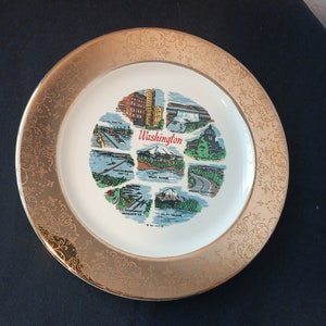 May include: A decorative plate with a gold rim featuring an ornate floral design. The center of the plate displays colorful illustrations of Washington state landmarks, including the text "Washington".