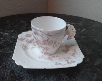 May include: A white porcelain teacup and saucer set with a floral pattern in shades of pink and gold. The cup has a unique, sculpted handle.