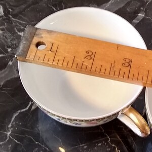 May include: White teacup with a gold handle and a decorative floral border. A wooden ruler is placed across the cup, measuring approximately 3 inches in diameter. The cup sits on a dark, marbled surface.