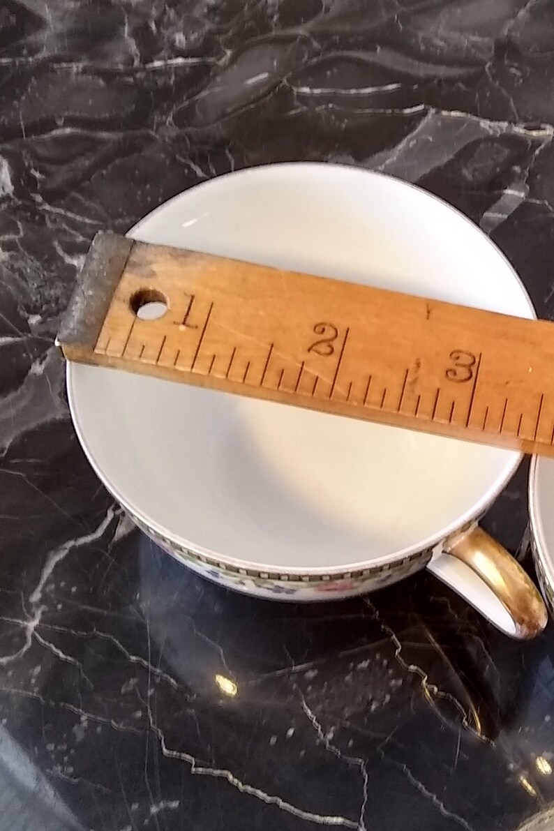 May include: White teacup with a gold handle and a decorative floral border. A wooden ruler is placed across the cup, measuring approximately 3 inches in diameter. The cup sits on a dark, marbled surface.
