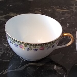 May include: White teacup with a gold handle and a floral design. The teacup has a green and gold border with pink and blue flowers. The teacup is on a black marble surface.