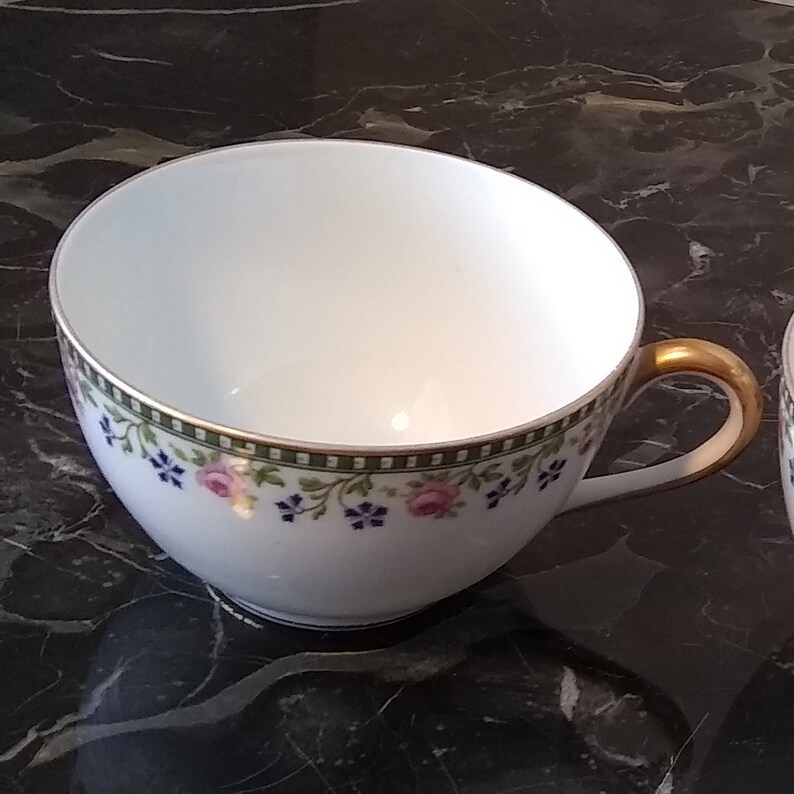 May include: White teacup with a gold handle and a floral design. The teacup has a green and gold border with pink and blue flowers. The teacup is on a black marble surface.