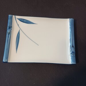 May include: Rectangular white ceramic dish with blue bamboo-style accents on the ends. The dish features a delicate blue leaf design. A stylish and elegant piece for serving appetizers or small dishes.