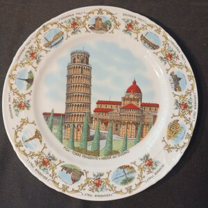 May include: A white ceramic plate with a gold trim and a colorful image of the Leaning Tower of Pisa and the Duomo in Pisa, Italy. The plate has the text "PISA - TORRE PENDENTE e ABSIDE del DUOMO" printed on it. The plate is surrounded by a border of small images of Italian landmarks and the text "SOUVENIR ITALY".