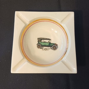 May include: White ceramic ashtray with a gold rim and a green and black illustration of a 1915 Ford Model T car. The text "Ford Model 1915" is printed below the car.