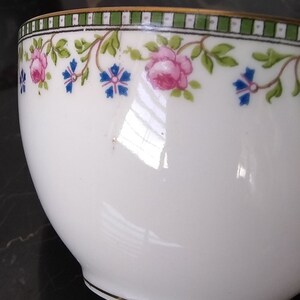 May include: White teacup with a floral design. The rim features a green and white striped border with pink roses and blue flowers. A thin gold band accents the rim and base of the cup. The cup has a glossy finish.
