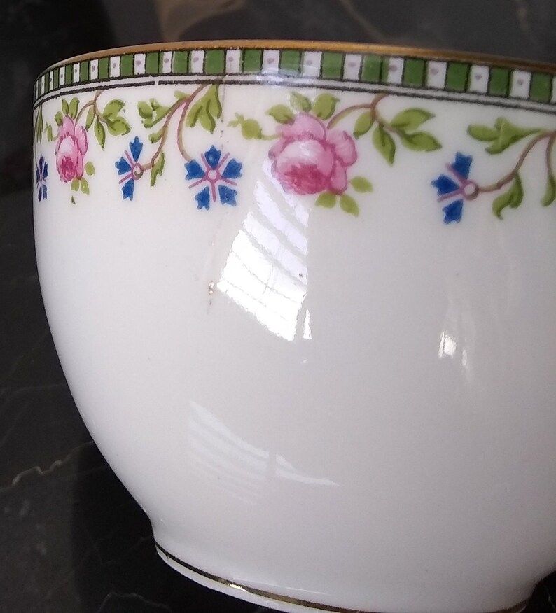May include: White teacup with a floral design. The rim features a green and white striped border with pink roses and blue flowers. A thin gold band accents the rim and base of the cup. The cup has a glossy finish.