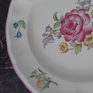 May include: A white ceramic plate with a pink, yellow, and blue floral design. The plate has a scalloped edge and a pink rim.