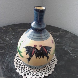 May include: A ceramic vase with a blue and white glaze, featuring a painted design of two black birds with red accents. The vase has a round body and a narrow neck.