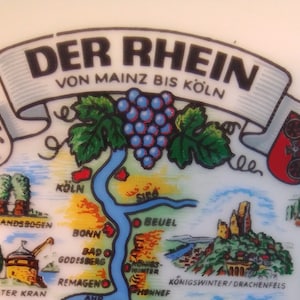 May include: A white ceramic plate with a colorful illustration of the Rhine River in Germany. The plate features a map of the river, with cities and landmarks marked along the way. The text "Der Rhein von Mainz bis Köln" is written in black lettering on a white banner.