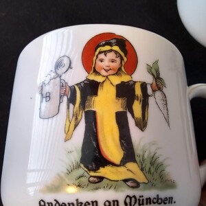 May include: White ceramic mug with a colorful illustration of a child holding a beer stein and a carrot. The text "Andenken an München." is printed on the bottom of the mug.