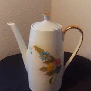 May include: White porcelain coffee pot with a gold trim and floral design. The flowers are blue, yellow, and pink.