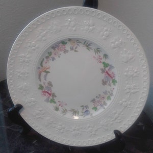 May include: White ceramic dinner plate with a raised embossed design around the edge and a floral pattern in shades of pink, yellow, blue, and green around the center.