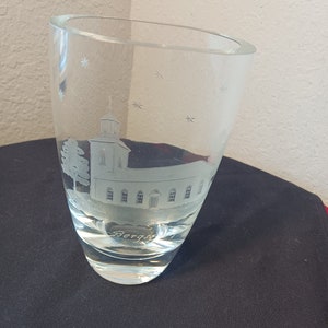 May include: A clear glass vase with an etched design of a church and the word "Bergs" at the bottom. The vase has a rounded bottom and a flared top.