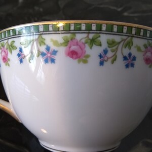 May include: White teacup with a floral design featuring pink roses and blue flowers. The rim has a green and white geometric pattern with a gold trim. The cup has a curved handle and a delicate appearance.