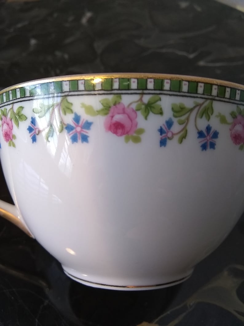 May include: White teacup with a floral design featuring pink roses and blue flowers. The rim has a green and white geometric pattern with a gold trim. The cup has a curved handle and a delicate appearance.