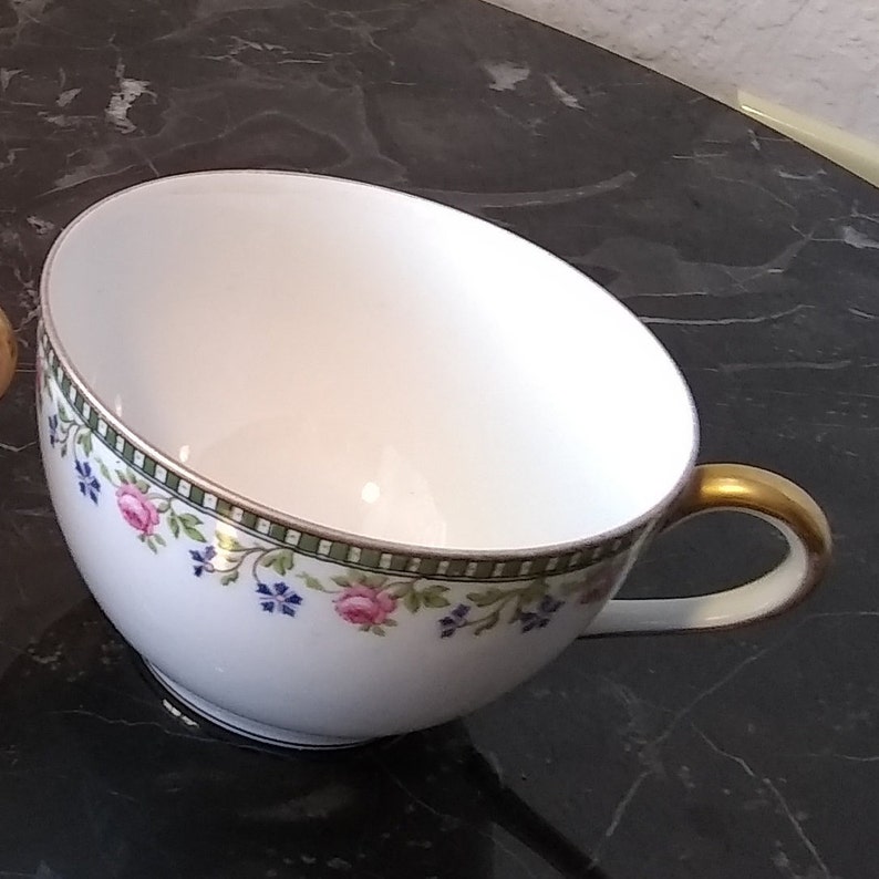 May include: White teacup with a gold handle and floral design. The cup features pink roses and blue flowers with green leaves, bordered by a green and gold band. The teacup is set against a dark background.