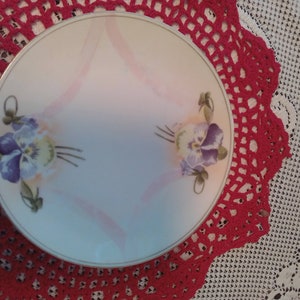 May include: A round, off-white decorative plate with a floral design featuring purple and yellow pansies. The plate has a gold rim and pink ribbon accents. The plate is set on a red crocheted doily.