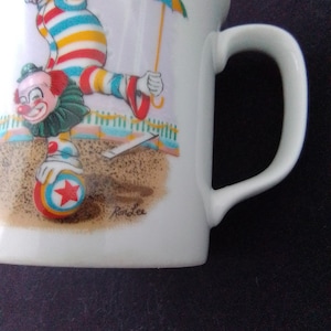 May include: White ceramic mug with a colorful illustration of a clown juggling a ball and holding an umbrella. The mug has a handle and the text "RonLee" is printed on the bottom.