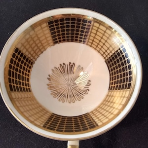 May include: A white porcelain teacup with a gold rim and a geometric pattern in black and gold. The cup has a stylized floral design in the center.