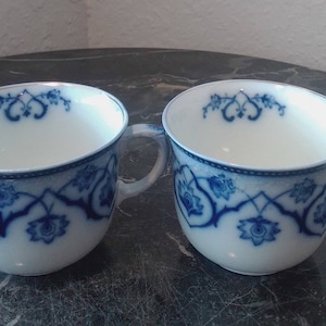 May include: Two white porcelain teacups with blue and gold floral designs. The cups have a rounded shape and a handle on the right side.