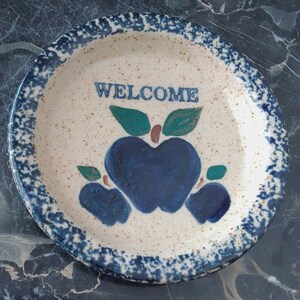 May include: A white ceramic plate with a blue and white speckled glaze. The plate has a blue and white apple design with the word "WELCOME" in the center.