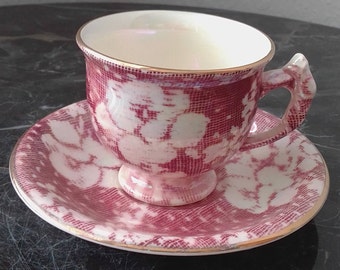 May include: A vintage teacup and saucer set with a pink and white floral pattern. The cup and saucer have a gold trim around the edges.