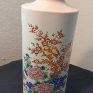 May include: A white ceramic vase with a pink glaze and a floral design. The vase features a branch of cherry blossoms with two yellow birds perched on the branches. The flowers are pink, white, and red, and the leaves are green. The vase has a gold rim.