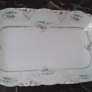 May include: A white rectangular serving platter with a scalloped edge and gold trim. The platter is decorated with pink and blue floral accents. The handles are curved. A decorative piece for serving food.