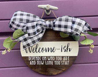 May include: A round wooden sign with a black and white checkered ribbon bow and greenery. The sign reads "Welcome-ish Depends on who you are and how long you stay."