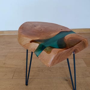 May include: A unique, handcrafted side table. The table features a live edge wood top with a teal resin river. The table is supported by three black hairpin legs. The table measures approximately 20 inches long, 18 inches wide, and 16 inches high.