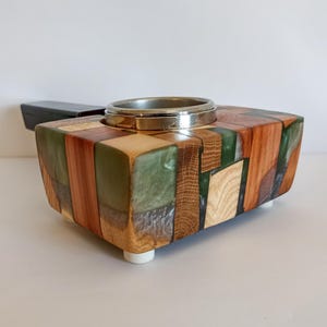 May include: A handcrafted wooden box with a silver ring insert. The box is made of various wood pieces in shades of brown, green, and natural wood tones. It has a rectangular shape and sits on four small white feet. The box is likely a decorative item.