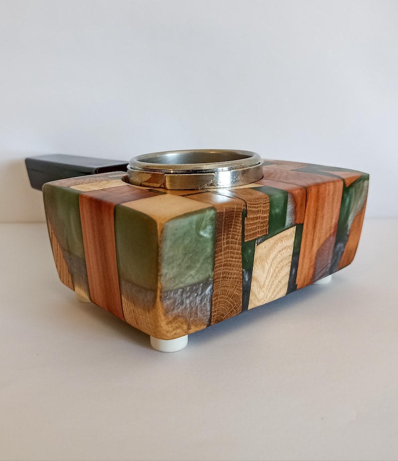 May include: A handcrafted wooden box with a silver ring insert. The box is made of various wood pieces in shades of brown, green, and natural wood tones. It has a rectangular shape and sits on four small white feet. The box is likely a decorative item.