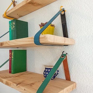 May include: Three natural wood shelves mounted to a wall with colorful leather straps. The top shelf has yellow straps, the middle has blue and black straps, and the bottom has green and tan straps. Books and small containers sit on the shelves.