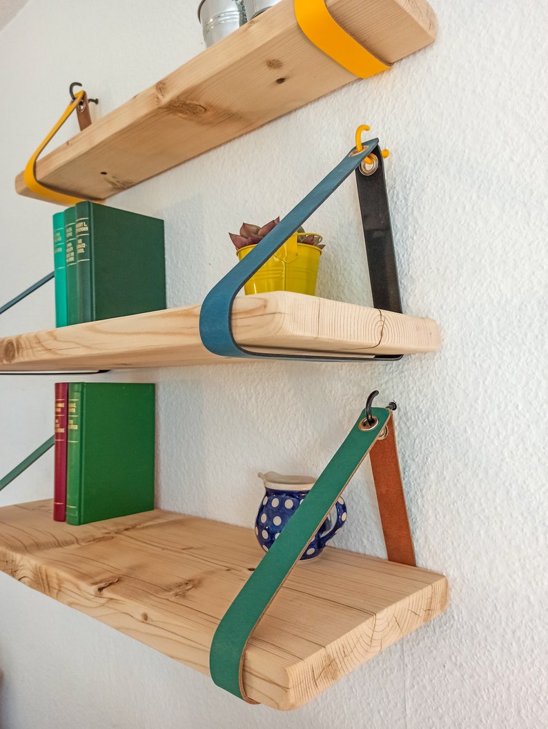 May include: Three natural wood shelves mounted to a wall with colorful leather straps. The top shelf has yellow straps, the middle has blue and black straps, and the bottom has green and tan straps. Books and small containers sit on the shelves.
