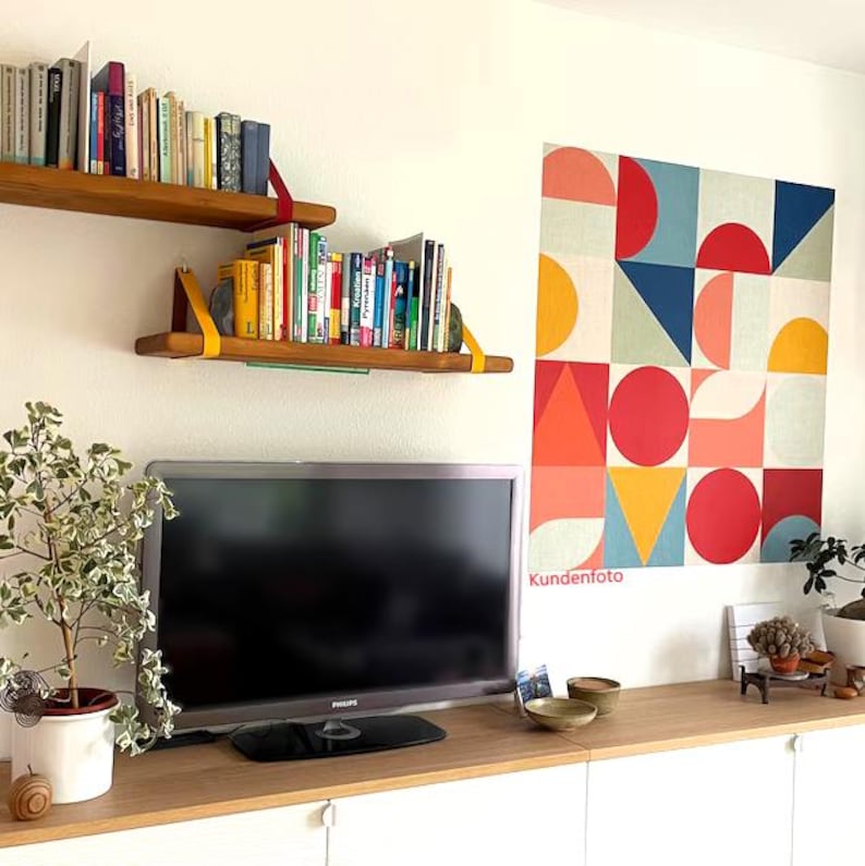 May include: A wall-mounted square canvas art piece featuring a geometric pattern of circles, triangles, and squares in red, orange, yellow, blue, and cream. The artwork is displayed above a wooden cabinet with a television and two floating shelves.
