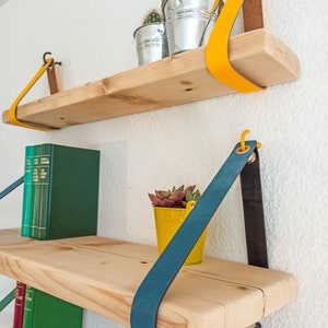 May include: Two light-colored wooden shelves suspended by colorful leather straps. The top shelf features a bright yellow strap and two silver metal buckets with cacti. The bottom shelf has a teal strap, a yellow bucket with a succulent, and green books.