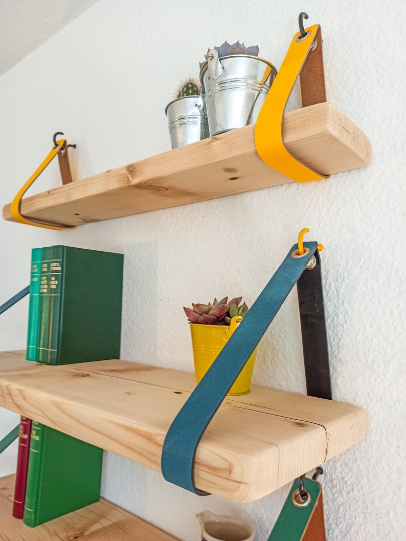 May include: Two light-colored wooden shelves suspended by colorful leather straps. The top shelf features a bright yellow strap and two silver metal buckets with cacti. The bottom shelf has a teal strap, a yellow bucket with a succulent, and green books.