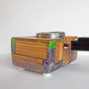 May include: A handcrafted wooden box with a silver metal top and a black handle. The box is made of various wood pieces, green resin, and purple resin. It has white feet.