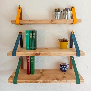 May include: Three light-colored wooden shelves suspended by leather straps in yellow, teal, and green. The top shelf holds two metal buckets with plants. The middle shelf features three green books and a yellow pot. The bottom shelf has two books and a blue polka-dot pitcher.