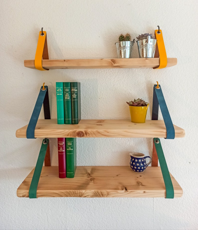 May include: Three light-colored wooden shelves suspended by leather straps in yellow, teal, and green. The top shelf holds two metal buckets with plants. The middle shelf features three green books and a yellow pot. The bottom shelf has two books and a blue polka-dot pitcher.
