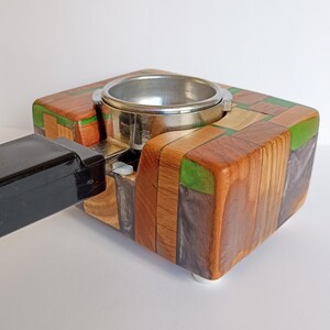 May include: A handcrafted espresso machine portafilter holder. The holder is made of various wood pieces in shades of brown, green, and tan, arranged in a mosaic pattern. A silver metal portafilter ring is set into the wood. A black handle is attached to the side.