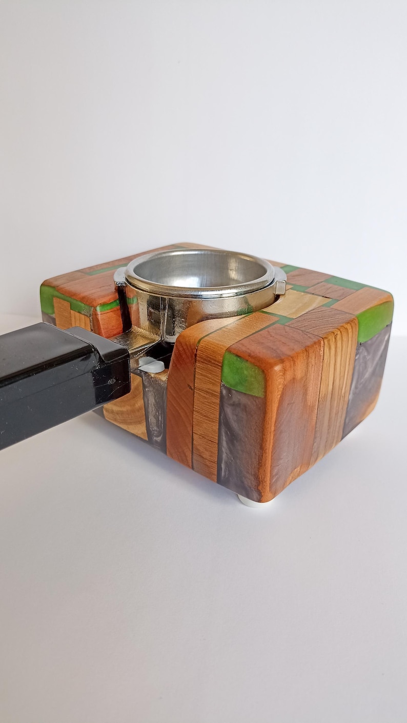 May include: A handcrafted espresso machine portafilter holder. The holder is made of various wood pieces in shades of brown, green, and tan, arranged in a mosaic pattern. A silver metal portafilter ring is set into the wood. A black handle is attached to the side.