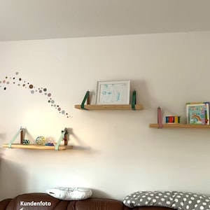 May include: Three light-colored wooden wall shelves suspended by leather straps in shades of mint green, dark green, and dusty pink. The shelves hold a white-framed picture, a colorful toy, and a children's book with a rainbow on the cover.