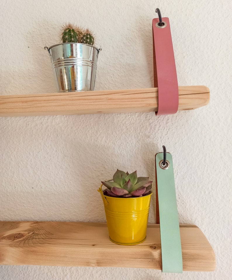 May include: Two horizontal light-colored wooden shelves mounted with vertical straps. The top shelf features a pink strap and a silver metal bucket with a cactus. The bottom shelf features a light green strap and a yellow metal bucket with a succulent.