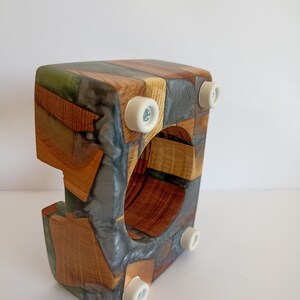 May include: A wooden wine bottle holder with a unique design. The holder is constructed from various wood pieces and resin, featuring a mix of natural wood tones and gray resin. It has a square shape with a circular opening and four white feet.