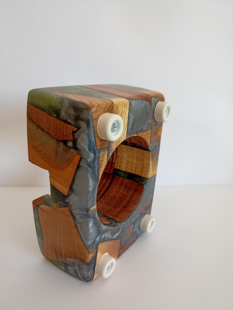 May include: A wooden wine bottle holder with a unique design. The holder is constructed from various wood pieces and resin, featuring a mix of natural wood tones and gray resin. It has a square shape with a circular opening and four white feet.