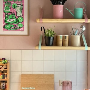 May include: Two light-colored wooden shelves suspended by leather straps. The top shelf holds a pink utensil holder and a mint green watering can. The bottom shelf holds a black pot with green sprouts, two cylindrical containers, and a beige utensil crock.