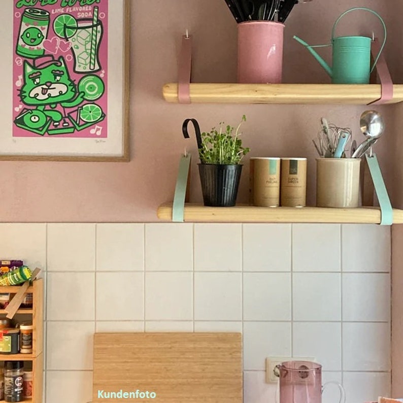 May include: Two light-colored wooden shelves suspended by leather straps. The top shelf holds a pink utensil holder and a mint green watering can. The bottom shelf holds a black pot with green sprouts, two cylindrical containers, and a beige utensil crock.