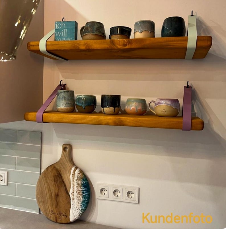 May include: Two horizontal wooden shelves mounted with light blue and purple leather straps. The top shelf holds a blue block with text and four ceramic mugs. The bottom shelf holds five ceramic mugs. A round wooden cutting board with blue resin.